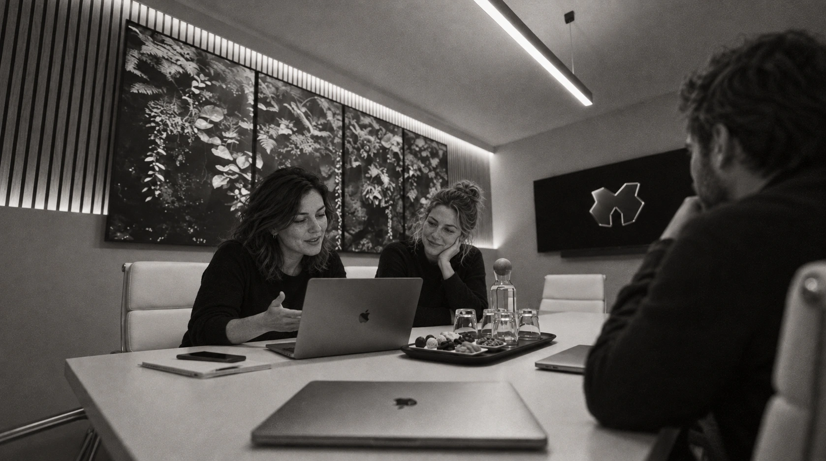 Three colleagues seated around the white table in Designco’s meeting room, discussing work on a MacBook Pro