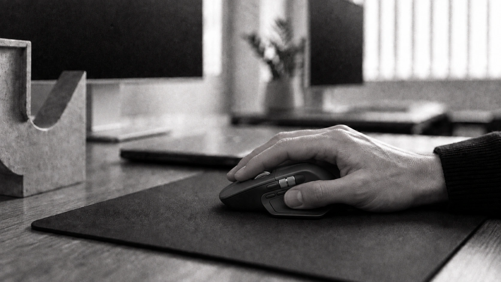 A hand using a graphite Logitech MX Master 4 mouse at a Designco workstation, with the studio softly out of focus