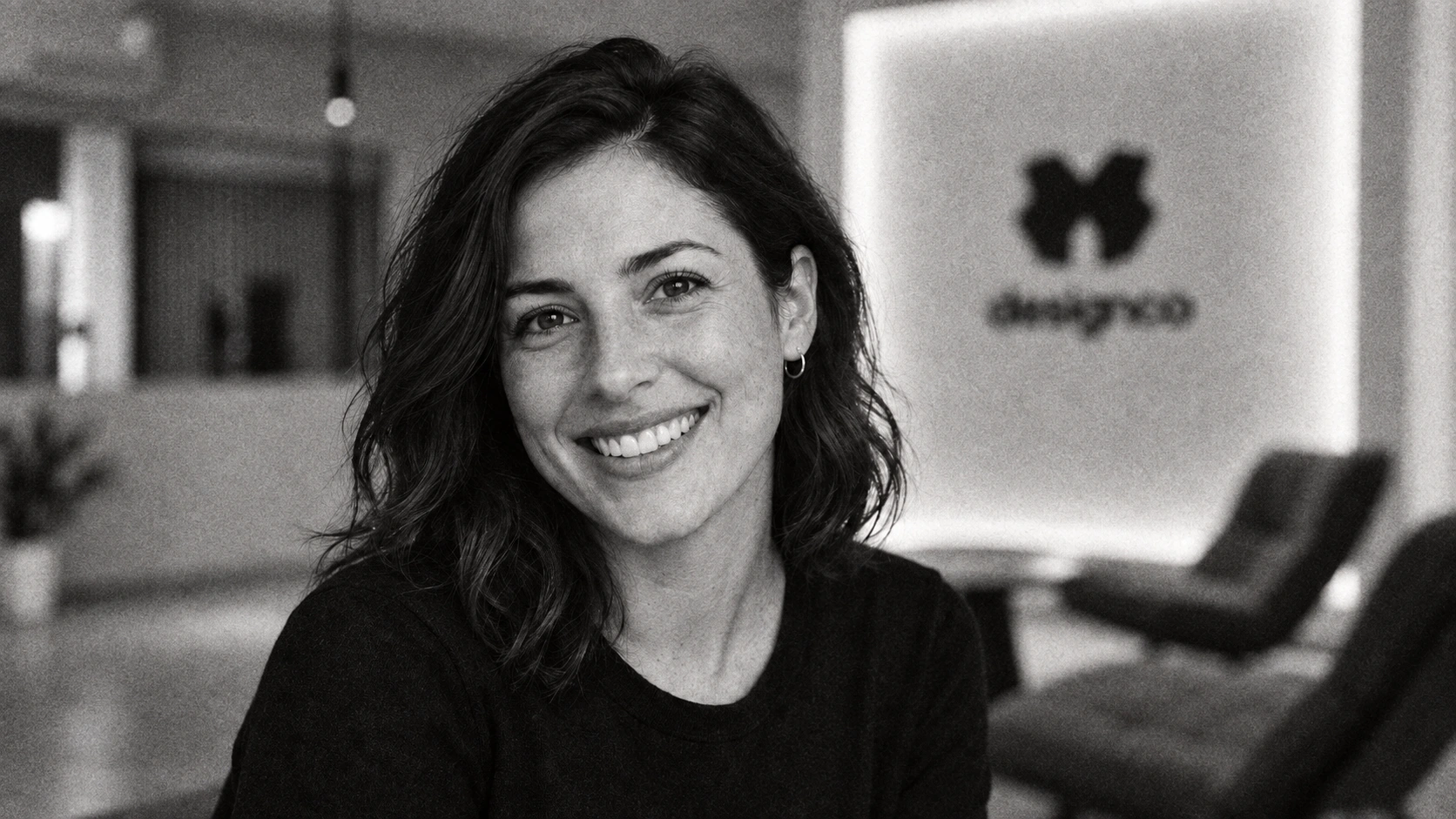 A smiling woman in a black shirt, with the Designco studio lounge softly blurred behind her
