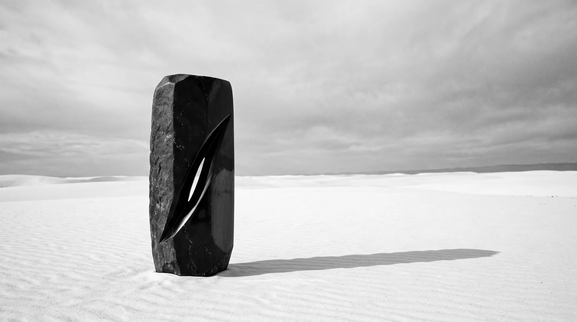 A single carved black obsidian form standing on a plain of white sand with a long shadow
